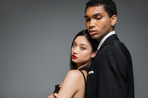 charming asian woman with red lips leaning on african american man in black blazer isolated on grey