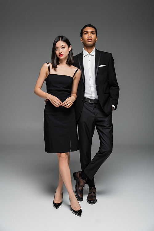 full length of asian woman in black dress and african american man in stylish suit standing on grey background