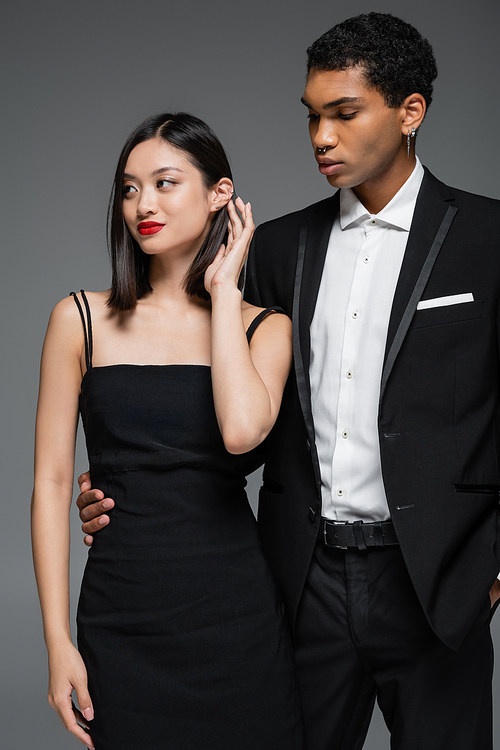 african american man in black blazer hugging waist of elegant asian woman smiling and looking away isolated on grey