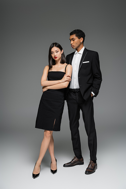 full length of asian woman in black dress and african american guy in elegant suit posing on grey background