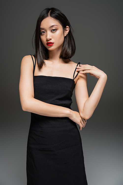 charming asian woman in black elegant dress looking at camera on grey background