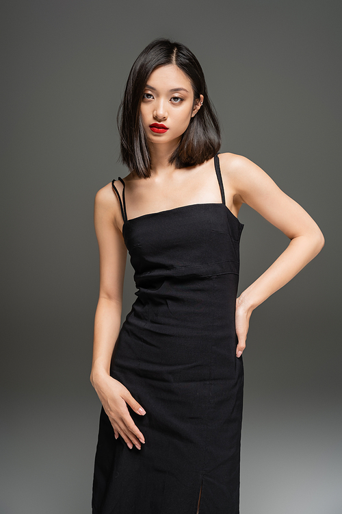 brunette asian woman in black stylish dress posing with hand on hip on grey background