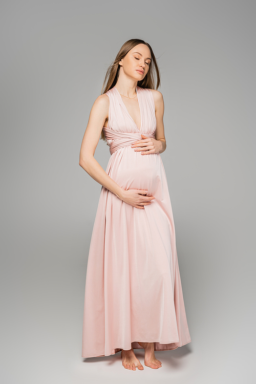 Full length of barefoot and fair haired pregnant woman in elegant pink dress touching belly and standing with closed eyes on grey background, elegant and stylish pregnancy attire