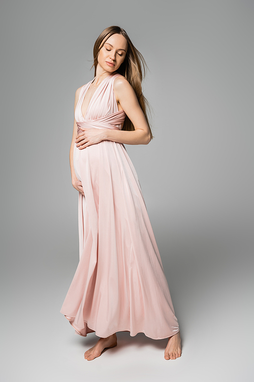 Full length of relaxed and barefoot pregnant woman in pink dress closing eyes while standing and posing on grey background, elegant and stylish pregnancy attire, sensuality, mother-to-be