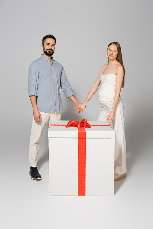 Smiling pregnant woman holding hand of husband and looking at camera while standing near big git box during gender reveal surprise party on grey background, expecting parents concept