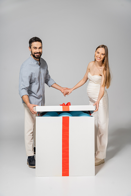 Cheerful and stylish couple holding hands while opening bog gift box with blue balloons during gender reveal surprise party on grey background, expecting parents concept, it`s a boy