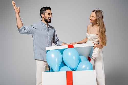 Excited bearded man looing at stylish pregnant wife opening big gift box with blue balloons during gender reveal surprise party and celebration isolated on grey, expecting parents concept, it`s a boy