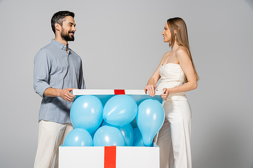 Cheerful and stylish pregnant woman looking at husband while opening big gift box with festive blue balloons during gender reveal surprise party isolated on grey, expecting parents, it`s a boy