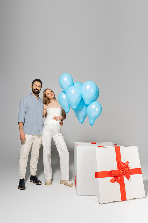 Full length of smiling and stylish expecting parents hugging while standing near big gift box with festive blue balloons during gender reveal surprise party on grey background, it`s a boy