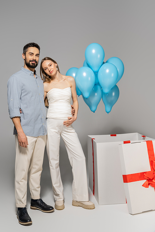 Full length of positive expecting parents hugging and looking at camera near big gift box with festive blue balloons during gender reveal surprise party on grey background, it`s a boy