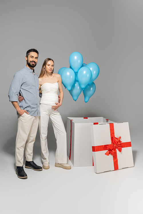 Full length of trendy and cheerful expecting parents hugging each other while standing near blue balloons and big gift box during gender reveal surprise party on grey background, it`s a boy