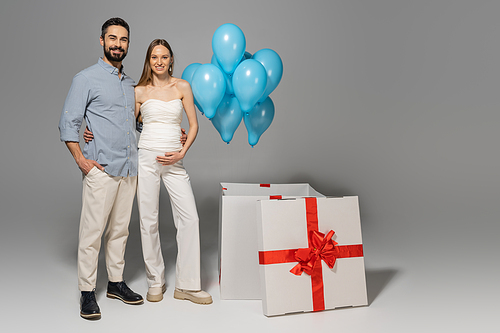 Full length of cheerful and trendy expecting parents hugging and looking at camera while standing near big gift box and festive blue balloons during gender reveal surprise party on grey background