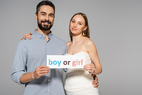 Positive and trendy pregnant woman holding card with boy or girl lettering and hugging stylish husband during gender reveal surprise party isolated on grey, expecting parents concept