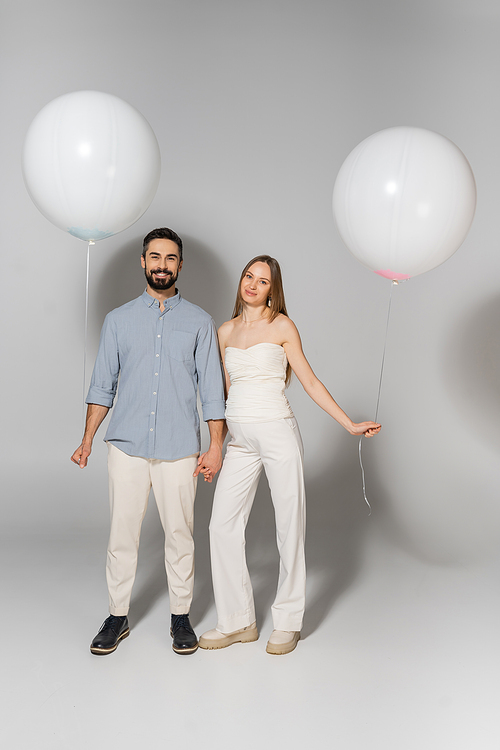 Full length of positive and stylish expecting parents looking at camera while holding hands and balloons during gender reveal surprise party on grey background, expecting parents concept