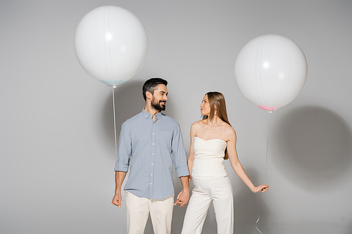 Positive and trendy expecting parents holding hands and festive balloons during celebration and gender reveal surprise party on grey background, expecting parents concept