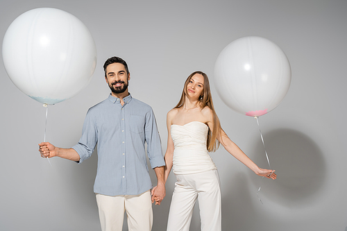 Smiling and stylish expecting parents holding hands and festive white balloons while looking at camera during gender reveal surprise party on grey background