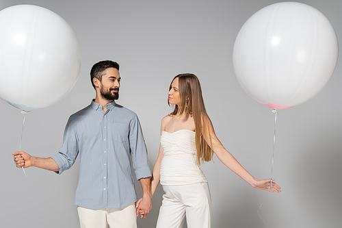 Smiling and trendy expecting parents holding hands and white festive balloons while looking at each other during celebration and gender reveal surprise party on grey background