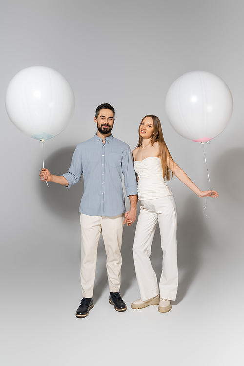 Full length of fashionable and happy expecting parents holding white balloons and looking at camera during gender reveal surprise party on grey background