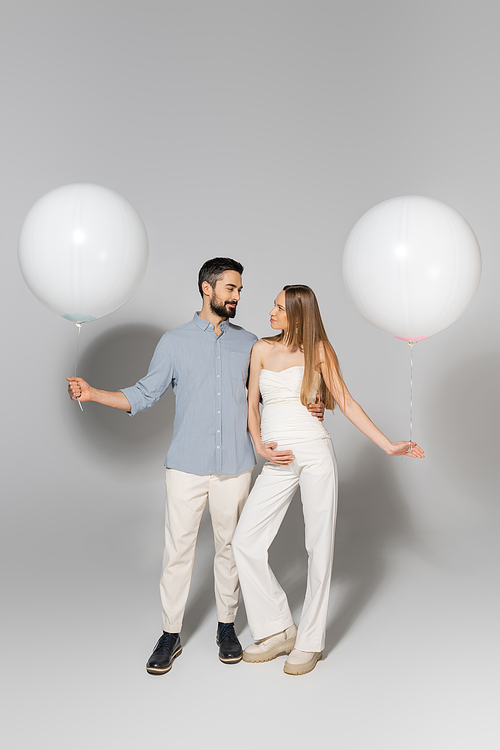 Full length of fashionable expecting parents hugging and looking at each other while holding festive balloons during gender reveal surprise party on grey background