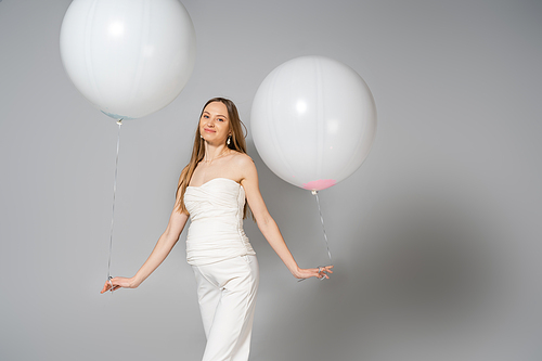 Positive and fashionable pregnant woman looking at camera while holding white festive balloons during gender reveal surprise party on grey background, fashionable pregnancy attire