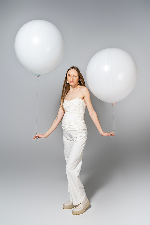 Full length of trendy and fair haired pregnant woman holding white balloons and looking at camera during gender reveal surprise party on grey background, fashionable pregnancy attire