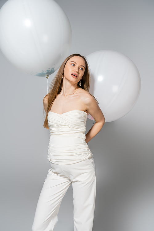 Pensive and fashionable pregnant woman looking away while holding festive white balloons and standing during gender reveal surprise party on grey background, fashionable pregnancy attire