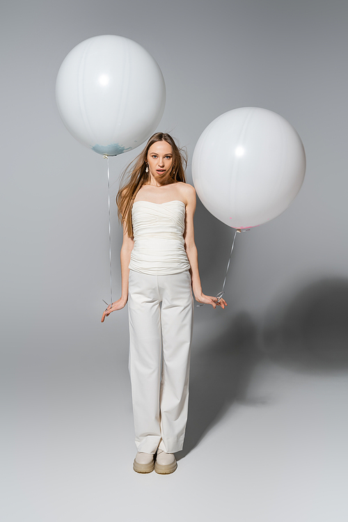 Full length of stylish and shocked expecting mother lookin at camera while holding white festive balloons during gender reveal surprise party on grey background, fashionable pregnancy attire