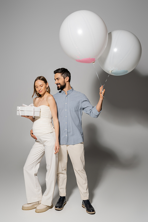 Cheerful man hugging stylish pregnant wife and holding gift box and festive balloons during celebration and gender reveal surprise party on grey background, fashionable pregnancy attire, boy or girl