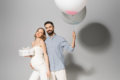 Positive and stylish man holding festive balloons and hugging pregnant wife with gift box during celebration and gender reveal surprise party on grey background, pregnancy attire, boy or girl