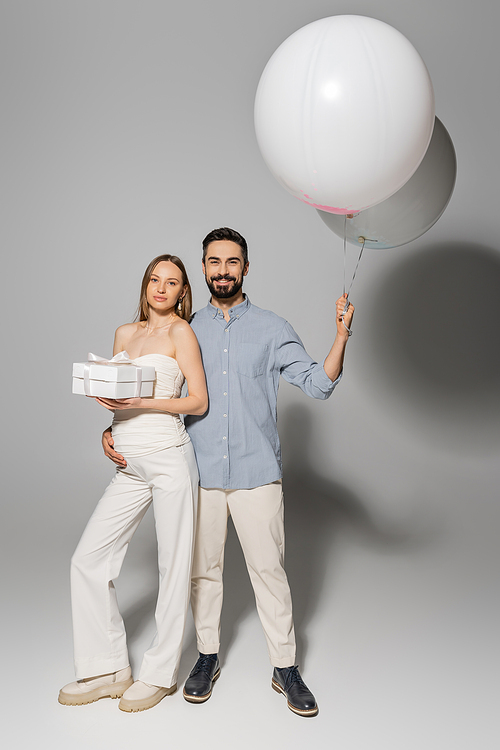 Full length of positive man holding festive balloons and hugging trendy and pregnant wife with gift box while standing together during baby shower party on grey background, boy or girl