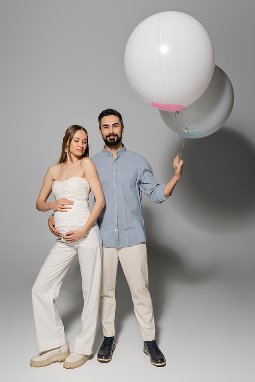 Full length of positive and stylish man holding festive balloons and hugging pregnant wife during baby shower party on grey background, expecting parents concept, boy or girl