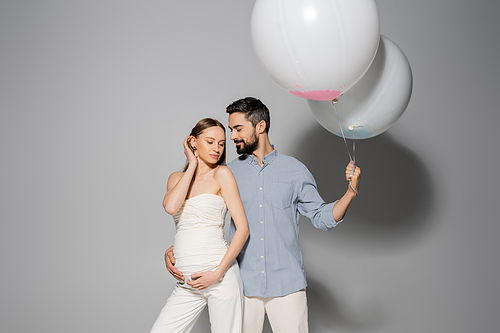 Smiling and bearded man hugging stylish pregnant wife and holding festive balloons during gender party and celebration on grey background, expecting parents concept, boy or girl