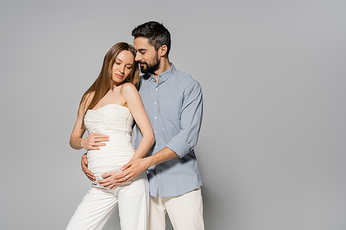 Trendy and bearded man hugging fair haired and pregnant woman while touching belly while standing together on grey background, expecting parents concept, baby bump, husband and wife