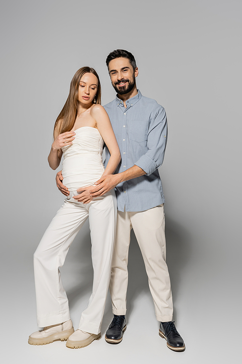 Full length of positive and bearded man hugging stylish and pregnant woman and looking at camera while standing together on grey background, expecting parents concept, baby bump, husband and wife