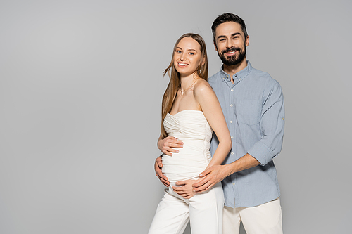 Smiling and bearded man hugging belly of fashionable pregnant woman and looking at camera while standing together isolated on grey, expecting parents concept, baby bump, husband and wife