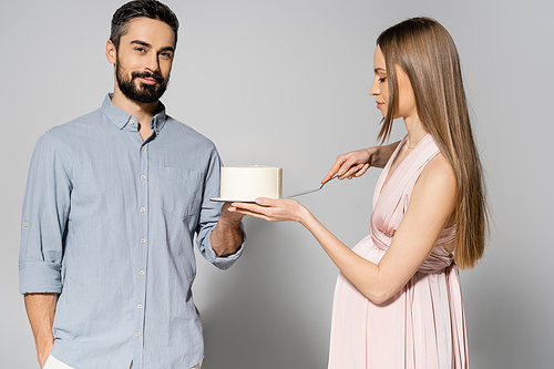 Stylish pregnant woman in dress cutting cake near smiling husband during baby shower party and celebration on grey background, expecting parents concept, gender party