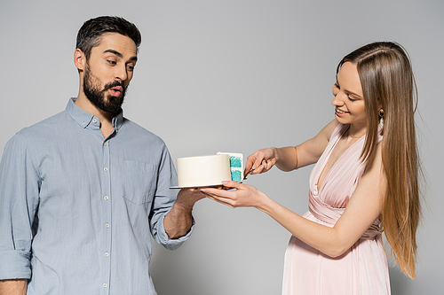 Smiling and elegant pregnant woman in pink dress cutting cake near shocked husband during baby shower party on grey background, expecting parents concept, gender party, it`s a boy