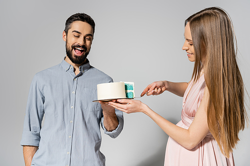 Excited and bearded man holding cake near cheerful pregnant wife with knife during baby shower party on grey background, expecting parents concept, gender party, it`s a boy