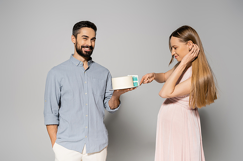 Positive and bearded man holding cake near elegant pregnant wife in pink dress during gender party on grey background, expecting parents concept, gender party, it`s a boy, cutting cake