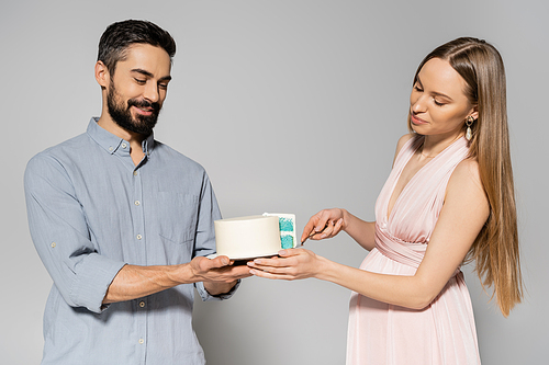 Elegant and pregnant woman in pink dress cutting blue cake near smiling husband during baby shower party on grey background, expecting parents concept, gender party, it`s a boy