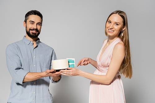 Cheerful and stylish couple looking at camera while cutting blue cake during baby shower celebration on grey background, expecting parents concept, gender party, it`s a boy