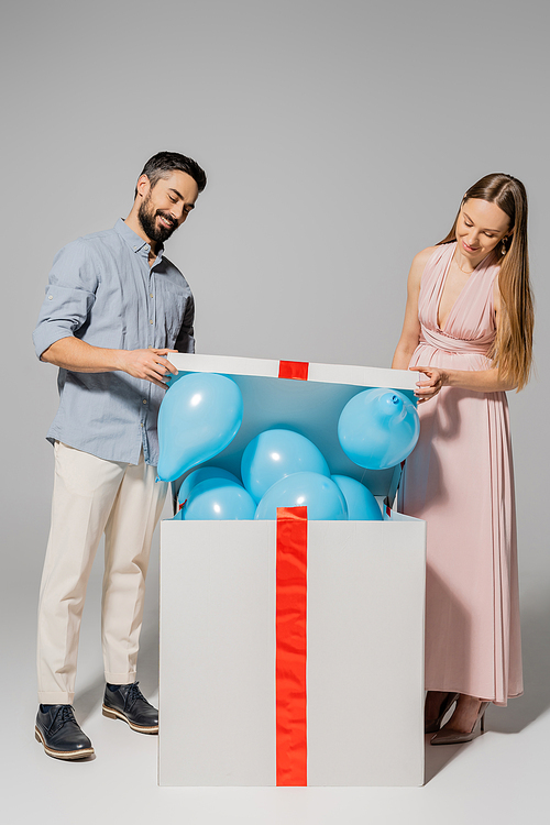 Positive and stylish couple opening big gift box with blue balloons during baby shower on grey background, expecting parents concept, gender party, it`s a boy