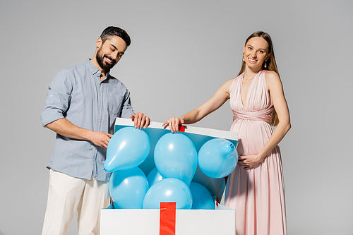 Smiling and elegant pregnant woman opening big gift box with blue balloons near husband during baby shower isolated on grey, expecting parents concept, gender party, it`s a boy