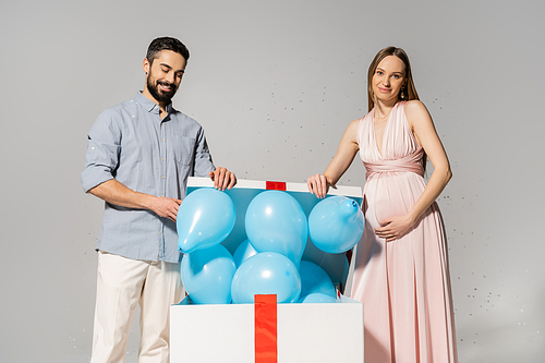 Positive and stylish couple opening gift box with blue balloons near festive confetti while celebrating baby shower party on grey background, expecting parents concept, gender party, it`s a boy