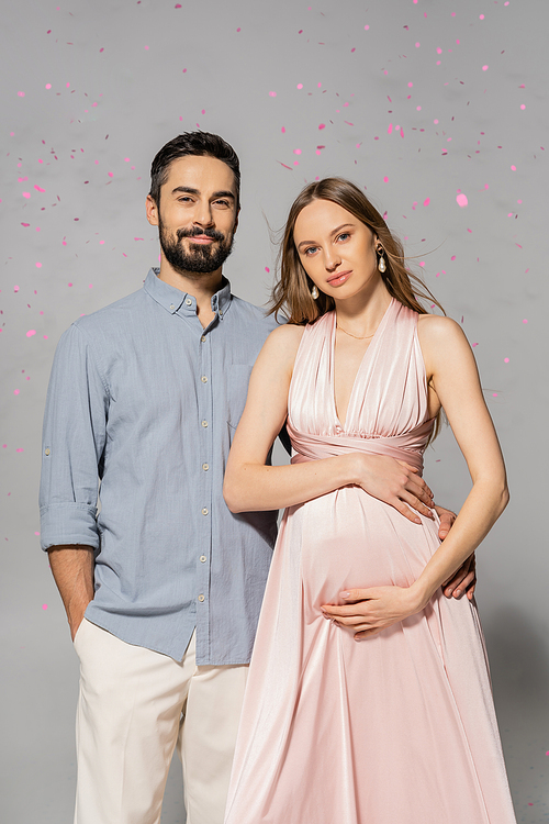 Portrait of smiling man hugging elegant and pregnant wife in pink dress while standing under confetti during baby shower party on grey background, expecting parents concept