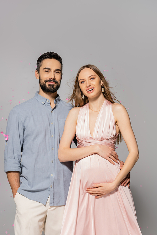 Positive man hugging elegant and pregnant wife in dress and looking at camera while standing under confetti during baby shower party on grey background, expecting parents concept