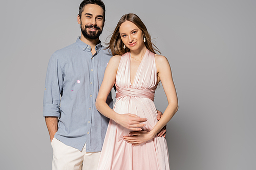 Stylish and cheerful man hugging elegant and pregnant wife while looking at camera and standing on grey background, expecting parents concept, new beginnings, parenthood