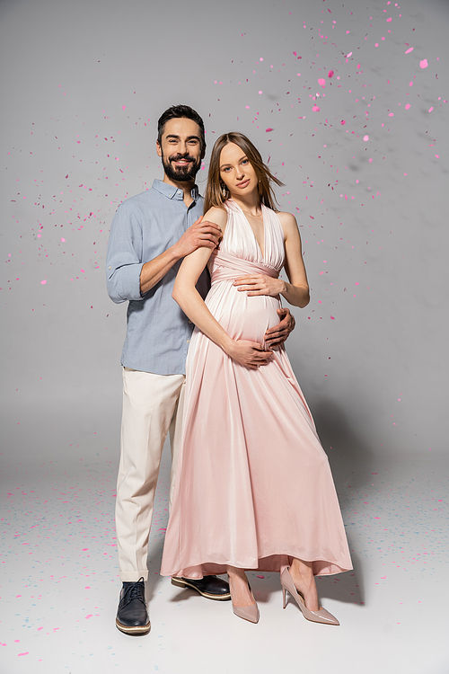 Full length of positive and elegant man hugging pregnant wife in dress while standing under confetti during baby shower party on grey background, expecting parents concept