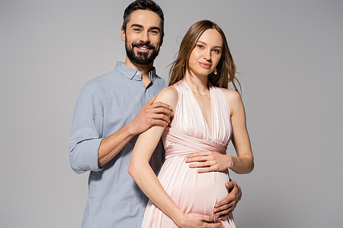 Portrait of positive and bearded man hugging elegant pregnant wife in pink dress while standing together isolated on grey, expecting parents concept, new beginnings, excitement