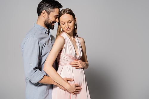 Happy and bearded man hugging pregnant woman in elegant dress while standing together on grey background, expecting parents concept, husband and wife